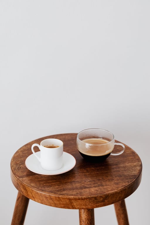 Fresh coffee in ceramic white cup and wide glass cup placed on round wooden table near white wall
